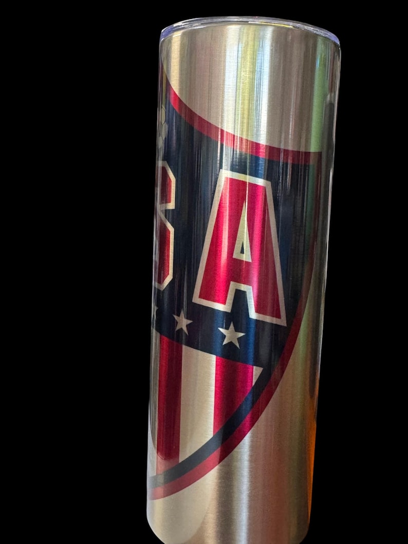 Puede incluir: Un vaso de acero inoxidable con un dise&ntilde;o patri&oacute;tico. El vaso presenta un escudo con las letras "USA" en rojo, blanco y azul, junto con estrellas y rayas. El dise&ntilde;o est&aacute; sobre un fondo plateado met&aacute;lico.