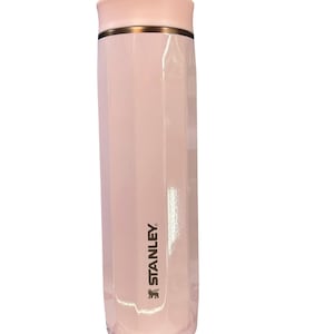 Peut inclure: Une gourde isotherme Stanley rose clair. Le bouchon est rose clair et orn&eacute; d'une bande dor&eacute;e. Le logo Stanley est imprim&eacute; en or sur le c&ocirc;t&eacute;. La gourde est cylindrique avec un design &agrave; facettes.