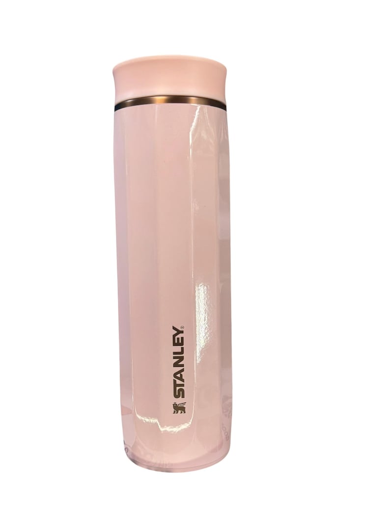 Peut inclure: Une gourde isotherme Stanley rose clair. Le bouchon est rose clair et orn&eacute; d'une bande dor&eacute;e. Le logo Stanley est imprim&eacute; en or sur le c&ocirc;t&eacute;. La gourde est cylindrique avec un design &agrave; facettes.