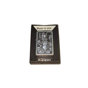 Puede incluir: Encendedor Zippo negro con un dise&ntilde;o steampunk que presenta a una mujer con gafas y engranajes. El encendedor est&aacute; en su caja original, con "Made in USA" impreso. El logotipo de Zippo es visible en la parte inferior.