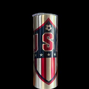 Puede incluir: Un vaso de acero inoxidable con un dise&ntilde;o patri&oacute;tico. El vaso presenta un escudo con las letras "USA" en rojo y blanco, un bal&oacute;n de f&uacute;tbol y estrellas. La mitad inferior del escudo tiene rayas rojas y blancas.