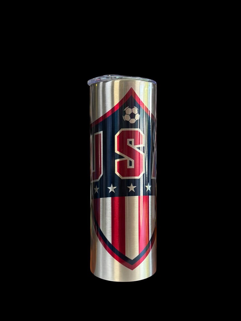 Puede incluir: Un vaso de acero inoxidable con un dise&ntilde;o patri&oacute;tico. El vaso presenta un escudo con las letras "USA" en rojo y blanco, un bal&oacute;n de f&uacute;tbol y estrellas. La mitad inferior del escudo tiene rayas rojas y blancas.