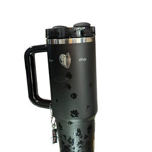 May include: Black insulated tumbler with a handle, featuring a floral design and the word "Ruby". The tumbler has a black lid and a silver rim. A small charm hangs from the handle.