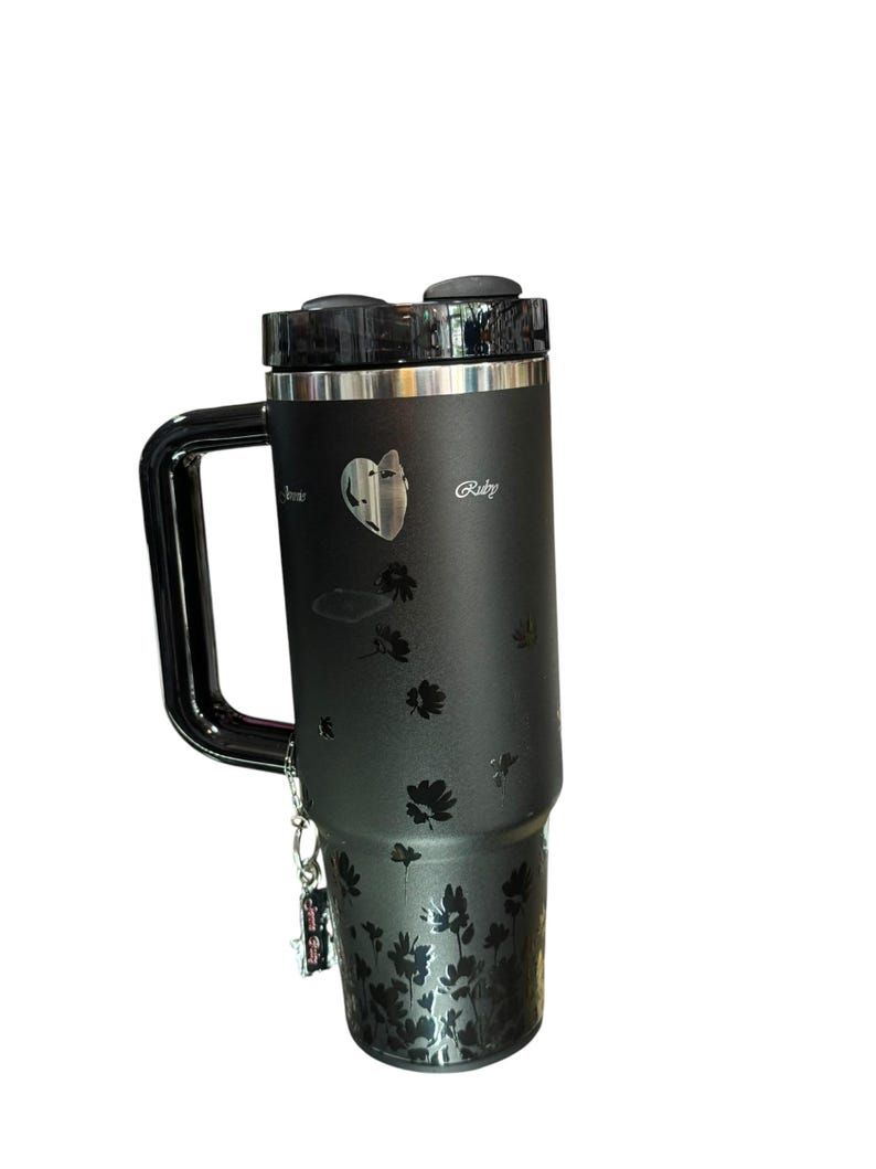 May include: Black insulated tumbler with a handle, featuring a floral design and the word "Ruby". The tumbler has a black lid and a silver rim. A small charm hangs from the handle.