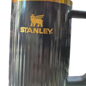 May include: A black Stanley mug with a gold rim and handle. The mug features the Stanley logo in gold, a winged bear, and the brand name. The mug has vertical ridges and a large handle.