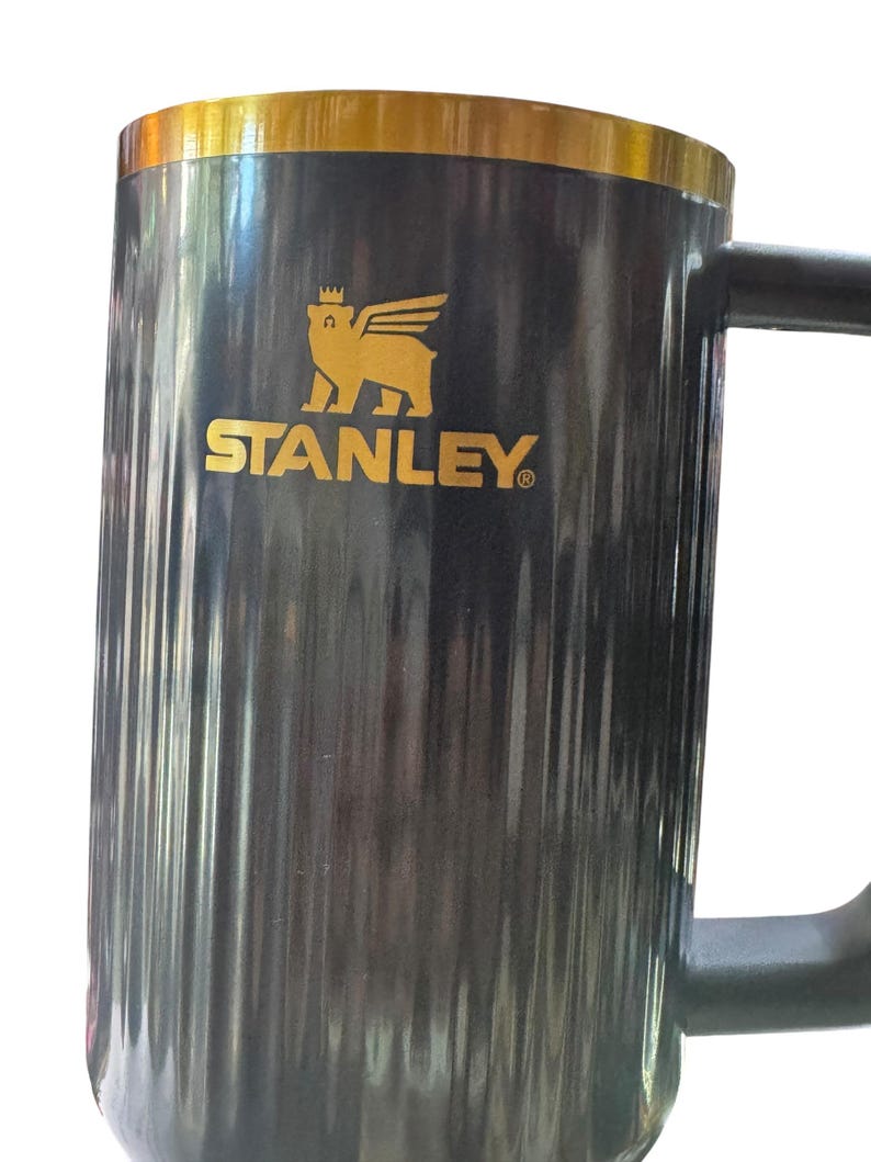 May include: A black Stanley mug with a gold rim and handle. The mug features the Stanley logo in gold, a winged bear, and the brand name. The mug has vertical ridges and a large handle.