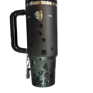 May include: Black travel mug with a handle, featuring a floral design and the name "Ruby". The mug has a silver rim and lid, and a small charm is attached to the handle. The mug is designed to keep beverages hot or cold.