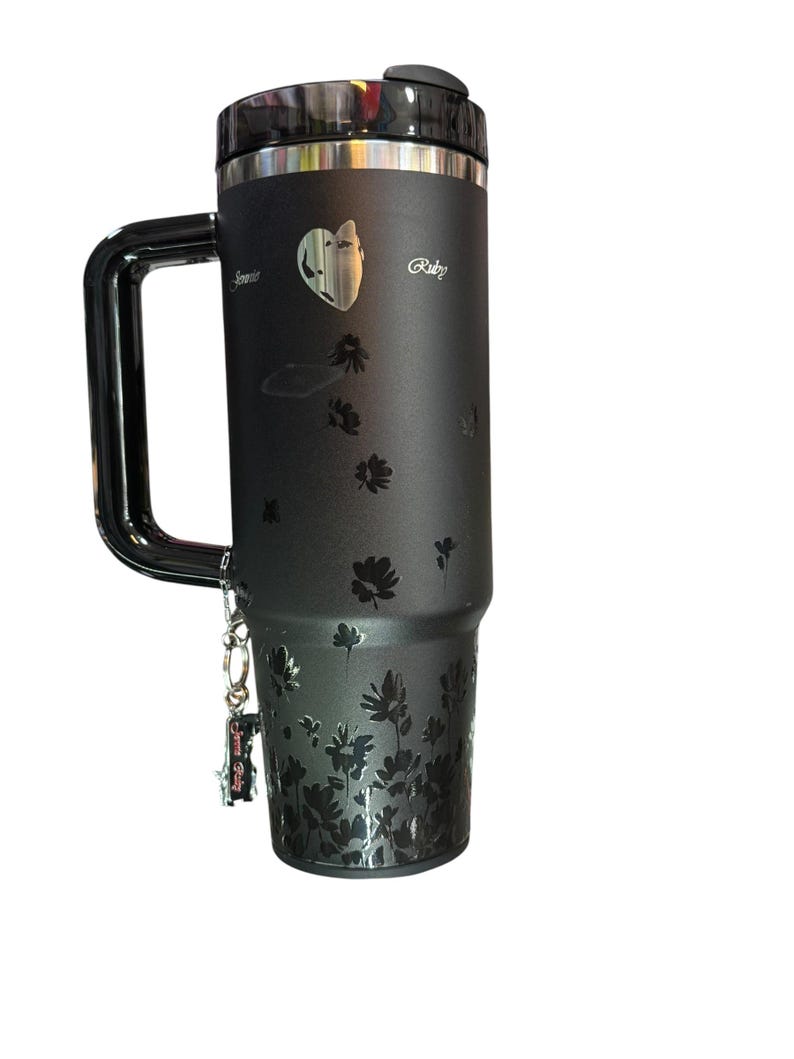 May include: Black travel mug with a handle, featuring a floral design and the name "Ruby". The mug has a silver rim and lid, and a small charm is attached to the handle. The mug is designed to keep beverages hot or cold.