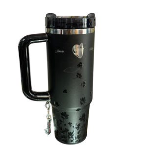 May include: Black travel mug with a handle, featuring a floral design and the name "Jennie". The mug has a silver rim and a black lid. A small charm hangs from the handle.