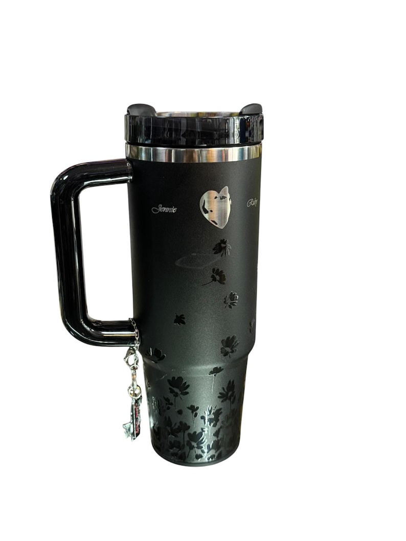 May include: Black travel mug with a handle, featuring a floral design and the name "Jennie". The mug has a silver rim and a black lid. A small charm hangs from the handle.