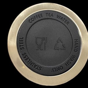 Puede incluir: Primer plano de una tapa de acero inoxidable con un centro negro. La tapa tiene las palabras "Coffee Tea Water" y "Stainless Steel" grabadas. Tambi&eacute;n tiene s&iacute;mbolos para lavar a mano solamente.