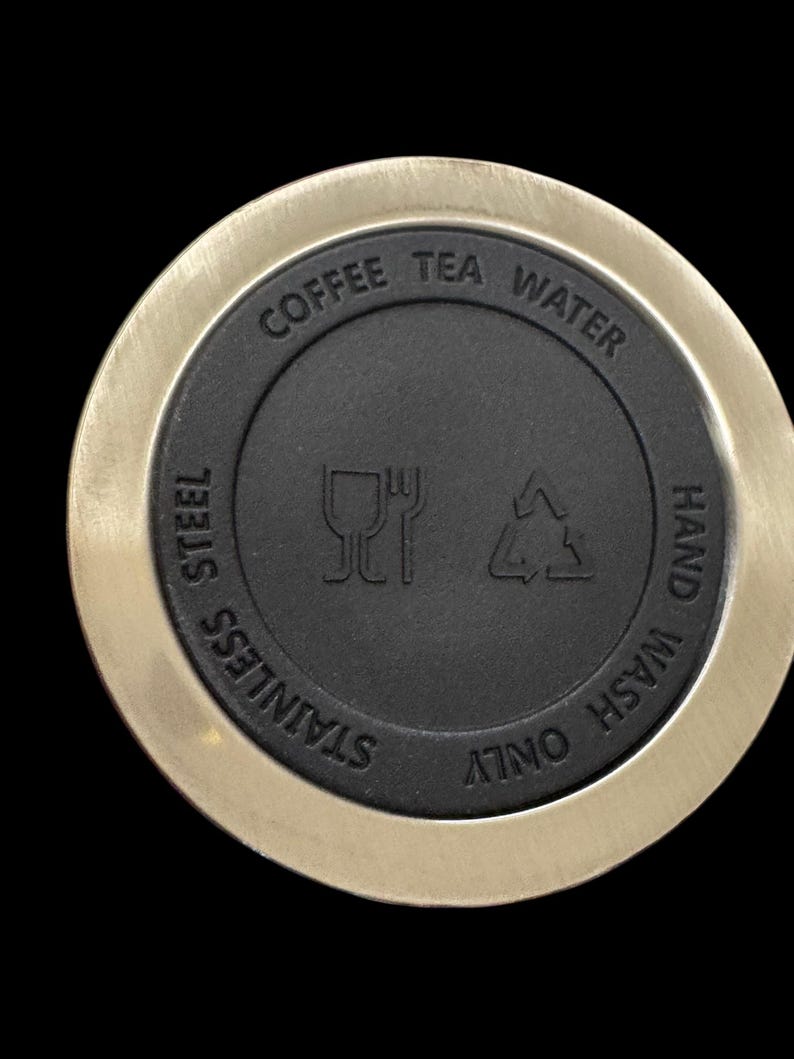 Puede incluir: Primer plano de una tapa de acero inoxidable con un centro negro. La tapa tiene las palabras "Coffee Tea Water" y "Stainless Steel" grabadas. Tambi&eacute;n tiene s&iacute;mbolos para lavar a mano solamente.
