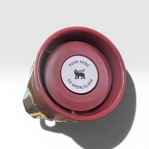 May include: A maroon travel mug with a push-button lid. The lid has a white sticker with the text "PUSH HERE TO OPEN/CLOSE" and a winged lion logo. The mug's exterior features a colorful, abstract design.
