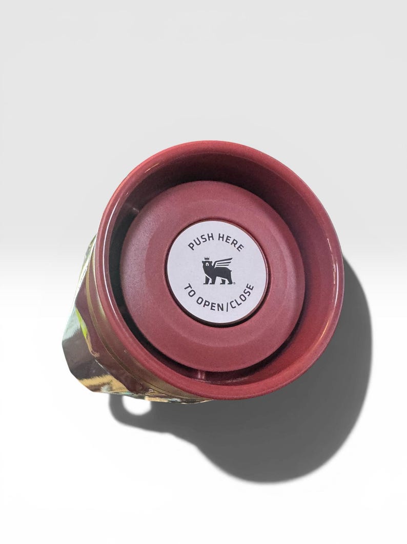May include: A maroon travel mug with a push-button lid. The lid has a white sticker with the text "PUSH HERE TO OPEN/CLOSE" and a winged lion logo. The mug's exterior features a colorful, abstract design.