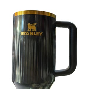 May include: Black and gold Stanley mug with a large handle. The mug has a gold rim and the Stanley logo in gold. The mug is cylindrical with vertical ridges. The mug is designed to keep beverages hot or cold.
