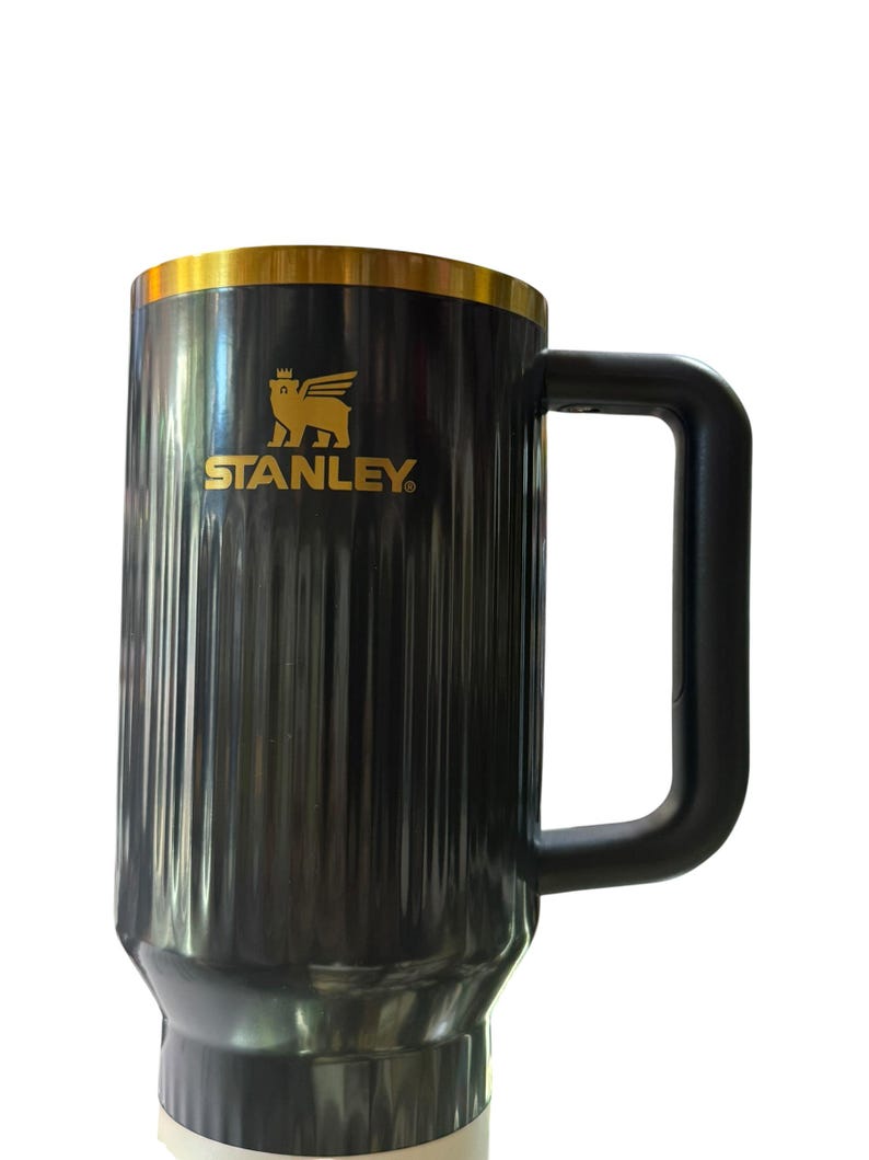 May include: Black and gold Stanley mug with a large handle. The mug has a gold rim and the Stanley logo in gold. The mug is cylindrical with vertical ridges. The mug is designed to keep beverages hot or cold.