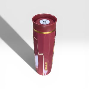 May include: A maroon Stanley thermos with a gold band and the Stanley logo in gold. The lid is maroon with a white sticker that says "PUSH HERE". The thermos is cylindrical and has a glossy finish.