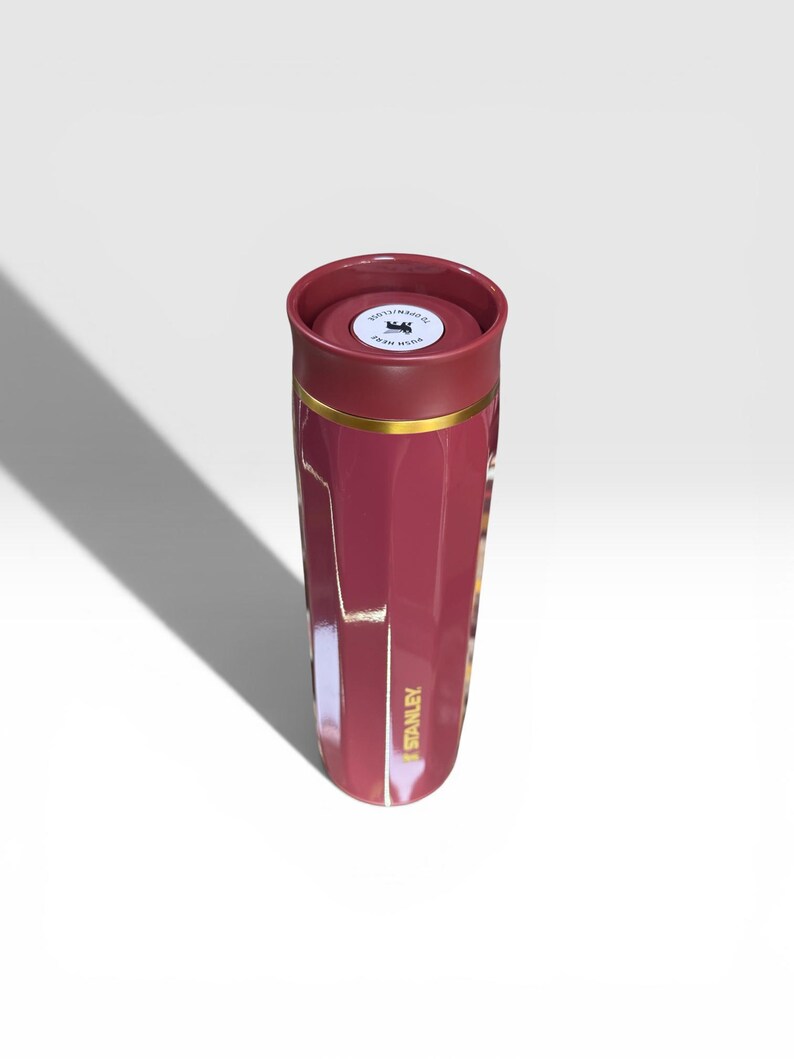 May include: A maroon Stanley thermos with a gold band and the Stanley logo in gold. The lid is maroon with a white sticker that says "PUSH HERE". The thermos is cylindrical and has a glossy finish.