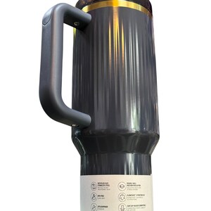 May include: A dark gray travel mug with a black handle and a gold-colored band near the top. The mug has a ridged design and a label with icons and text. The mug is made of stainless steel and is dishwasher safe.