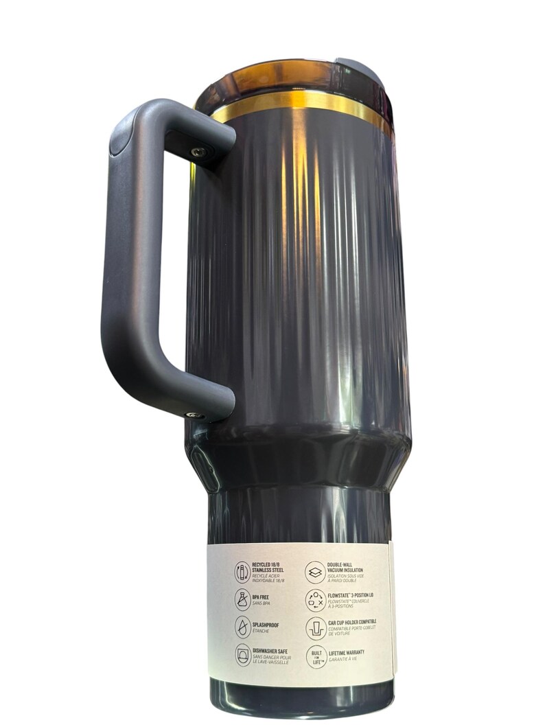 May include: A dark gray travel mug with a black handle and a gold-colored band near the top. The mug has a ridged design and a label with icons and text. The mug is made of stainless steel and is dishwasher safe.