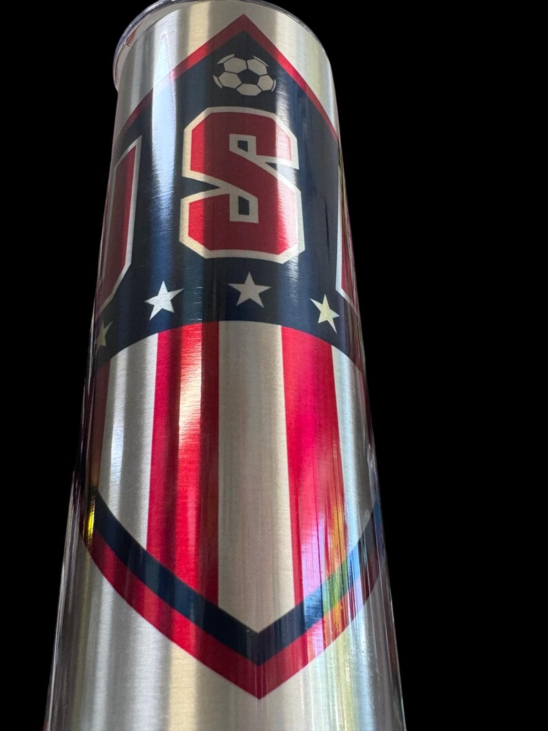 Puede incluir: Un vaso de acero inoxidable con un dise&ntilde;o patri&oacute;tico. El vaso presenta una combinaci&oacute;n de colores rojo, blanco y azul con las letras "USA" en rojo, un gr&aacute;fico de bal&oacute;n de f&uacute;tbol y rayas verticales rojas y blancas. El dise&ntilde;o est&aacute; sobre un fondo plateado.