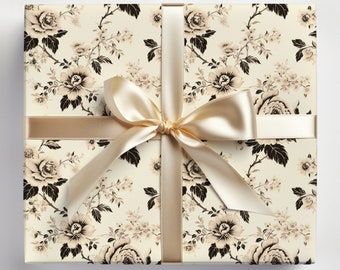 Puede incluir: Una caja de regalo envuelta en papel crema con un estampado floral negro y marrón. Una cinta de satén dorada está atada en un lazo en la parte superior de la caja.