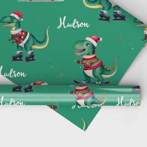 May include: Green wrapping paper with a repeating pattern of dinosaurs wearing Santa hats and roller skating. The dinosaurs are green with red and white sweaters. The text "Hudson" is repeated on the paper.