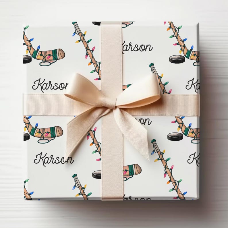 K&ouml;nnte beinhalten: Wei&szlig;e Geschenkpapier mit einem sich wiederholenden Muster aus Hockeyschl&auml;gern mit Weihnachtslichtern und dem aufgedruckten Namen "Karson". Das Geschenkpapier ist mit einem beigefarbenen Band gebunden.