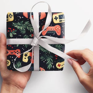 K&ouml;nnte beinhalten: Ein Geschenk, verpackt mit einem schwarzen Hintergrund, der mit roten und gelben Gamecontrollern, gr&uuml;nen Zweigen und wei&szlig;en Punkten gemustert ist. Das Geschenk ist mit einem wei&szlig;en Band und einer Schleife gebunden.