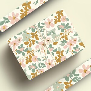 Op de afbeelding: Een cadeau verpakt in wit bloemmotiefpapier met een roze, groene en gouden bloemenpatroon. Het papier is vastgebonden met touw en de uiteinden van het touw zijn vastgebonden in een strik.
