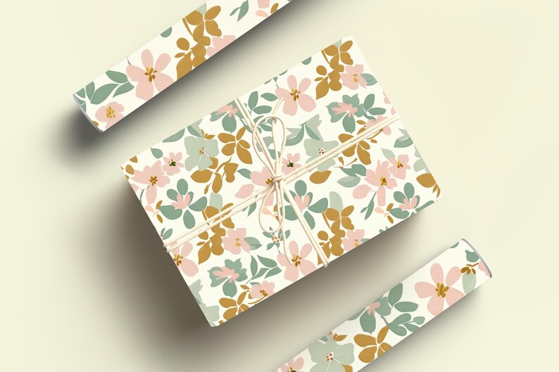 Op de afbeelding: Een cadeau verpakt in wit bloemmotiefpapier met een roze, groene en gouden bloemenpatroon. Het papier is vastgebonden met touw en de uiteinden van het touw zijn vastgebonden in een strik.