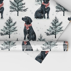 K&ouml;nnte beinhalten: Geschenkpapier mit einem sich wiederholenden Muster aus schwarzen Labrador Retrievern, die rot-wei&szlig; karierte Schals tragen, vor dem Hintergrund stilisierter immergr&uuml;ner B&auml;ume und Schneeflocken auf wei&szlig;em Grund. Eine Rolle des gleichen Papiers ist ebenfalls sichtbar.