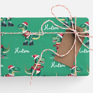 May include: Green wrapping paper with a repeating pattern of dinosaurs wearing Santa hats and ice skating. The paper is tied with red and white twine and has a brown gift tag with the name "Hudson" written on it.