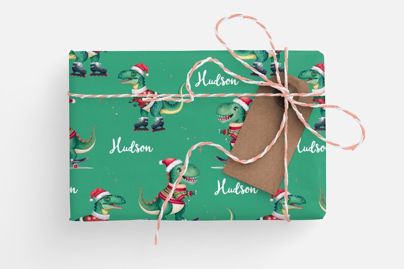May include: Green wrapping paper with a repeating pattern of dinosaurs wearing Santa hats and ice skating. The paper is tied with red and white twine and has a brown gift tag with the name "Hudson" written on it.