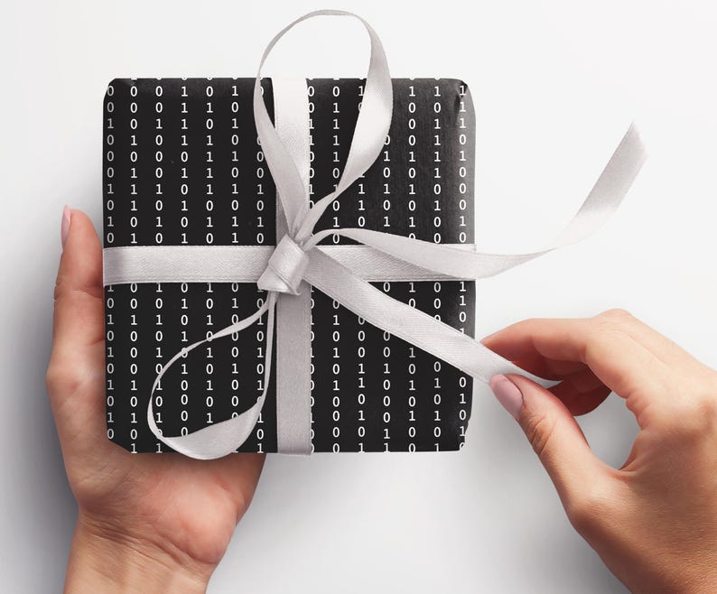 May include: A black gift box with a white ribbon and a pattern of 0s and 1s. The box is being held by a person's hands.