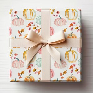 May include: A wrapped gift box featuring a white background with a repeating pattern of watercolor pumpkins in shades of orange, pink, and green, along with autumn leaves. The box is tied with a beige ribbon and bow, creating a festive presentation.