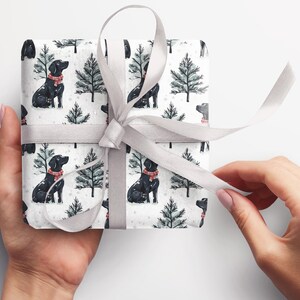 K&ouml;nnte beinhalten: Ein verpacktes Geschenk mit wei&szlig;em Hintergrund. Das Geschenkpapier zeigt ein Muster aus schwarzen Hunden mit roten Schals und gr&uuml;nen Weihnachtsb&auml;umen auf wei&szlig;em Grund. Ein silbernes Band ist um das Geschenk gebunden.