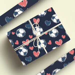 K&ouml;nnte beinhalten: Zwei Rollen Geschenkpapier mit dunkelblauem Hintergrund und einem Muster aus wei&szlig;en Fu&szlig;b&auml;llen und rosa Herzen.