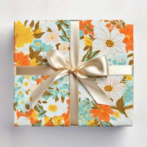 May include: A gift box wrapped in floral patterned paper with a light brown satin ribbon tied in a bow. The paper features a variety of flowers in shades of orange, yellow, blue, and white on a light blue background.