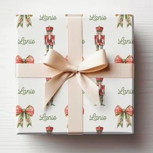 May include: A gift box wrapped in white paper, patterned with nutcrackers, bows, and banners in red, green, and cream. A cream ribbon and bow adorn the box. The name "Lanie" is written in a script font across the paper.
