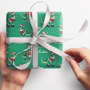 May include: Green wrapping paper with a repeating pattern of dinosaurs wearing Santa hats and riding skateboards. The paper is tied with a white ribbon and the name "Hudson" is written on the paper.