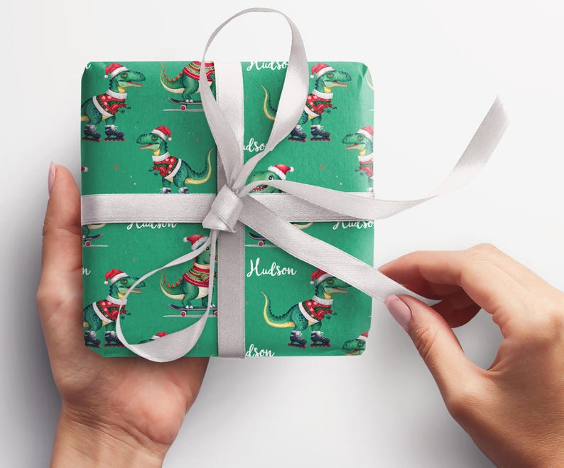 May include: Green wrapping paper with a repeating pattern of dinosaurs wearing Santa hats and riding skateboards. The paper is tied with a white ribbon and the name "Hudson" is written on the paper.
