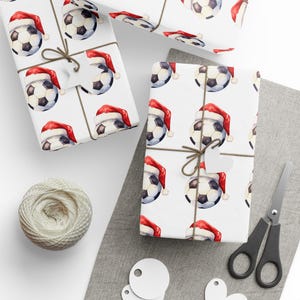 May include: White wrapping paper with a repeating pattern of soccer balls wearing Santa hats. The paper is tied with twine and a white gift tag.