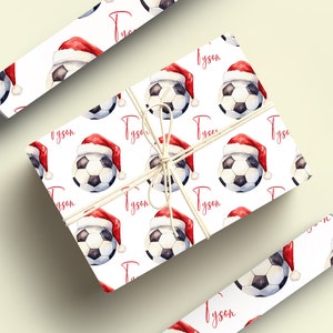 May include: White wrapping paper with a repeating pattern of soccer balls wearing Santa hats. The name "Tyson" is printed in red on the paper.