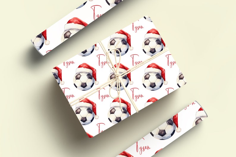 May include: White wrapping paper with a repeating pattern of soccer balls wearing Santa hats. The name "Tyson" is printed in red on the paper.