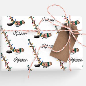 K&ouml;nnte beinhalten: Wei&szlig;es Geschenkpapier mit einem sich wiederholenden Muster aus Hockeyschl&auml;gern mit Weihnachtslichtern und dem Namen "Karson" auf jedem Schl&auml;ger. Das Papier ist mit einem braunen und wei&szlig;en Bindfaden gebunden.