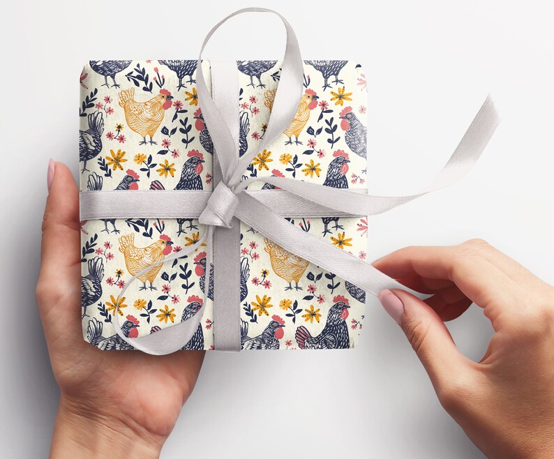 Puede incluir: Un papel de regalo blanco con un patr&oacute;n repetitivo de pollos negros y amarillos y flores. El regalo est&aacute; atado con una cinta de sat&eacute;n blanca.
