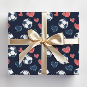 K&ouml;nnte beinhalten: Eine dunkelblaue Geschenkpapier mit einem Muster aus wei&szlig;en Fu&szlig;b&auml;llen und rosa Herzen. Ein goldenes Band ist um die Schachtel gebunden.