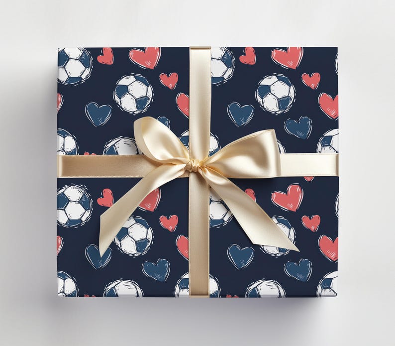 K&ouml;nnte beinhalten: Eine dunkelblaue Geschenkpapier mit einem Muster aus wei&szlig;en Fu&szlig;b&auml;llen und rosa Herzen. Ein goldenes Band ist um die Schachtel gebunden.