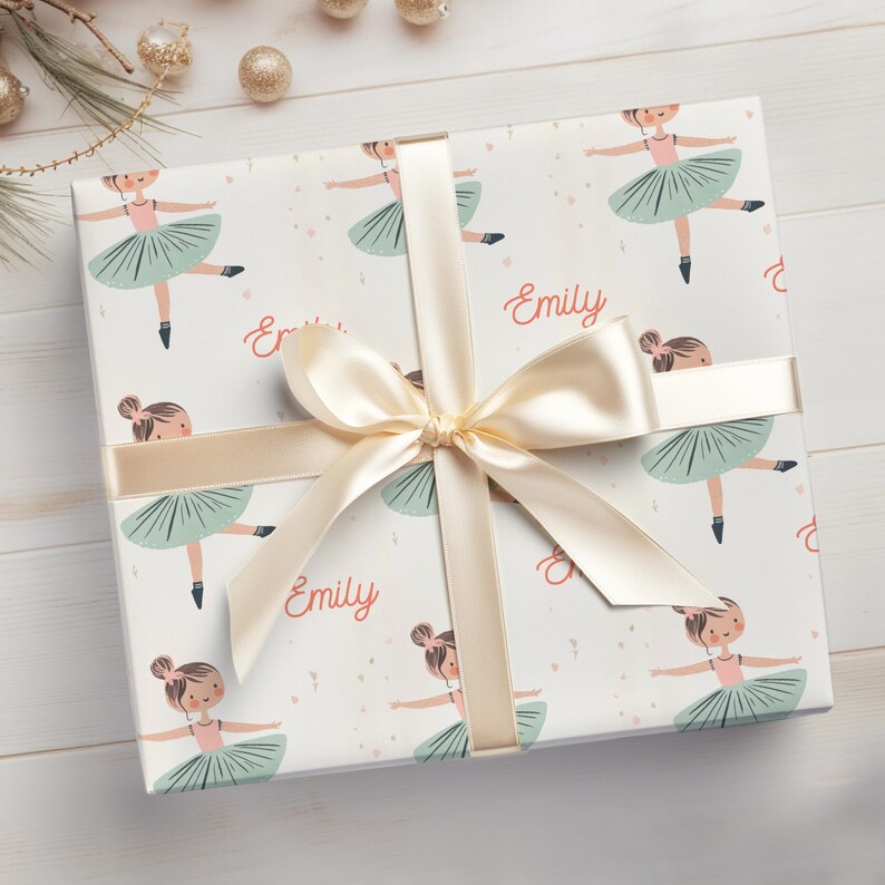 May include: White gift wrap with a repeating pattern of ballerina girls in pink and green tutus. The wrapping paper is tied with a cream-colored ribbon and bow. The name "Emily" is printed on the wrapping paper.